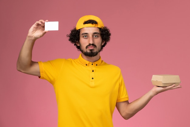 Front view male courier in yellow uniform and cape with card and little delivery food package on his hands on the light pink background.