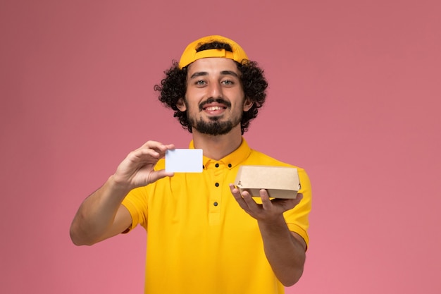밝은 분홍색 배경에 그의 손에 카드와 작은 배달 음식 패키지와 노란색 유니폼과 케이프에서 전면보기 남성 택배.