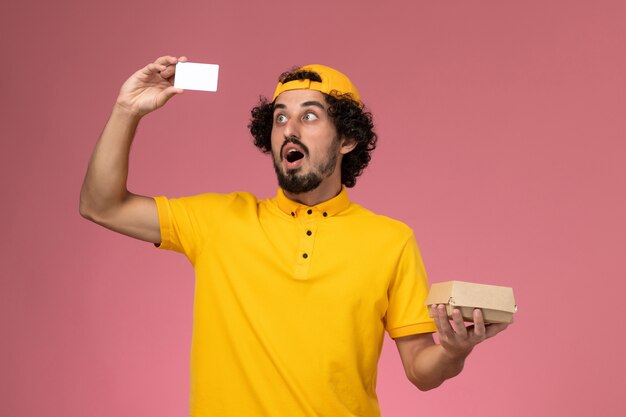 Вид спереди мужской курьер в желтой форме и плащ с карточкой и небольшой пакет продуктов для доставки на руках на светло-розовом фоне.