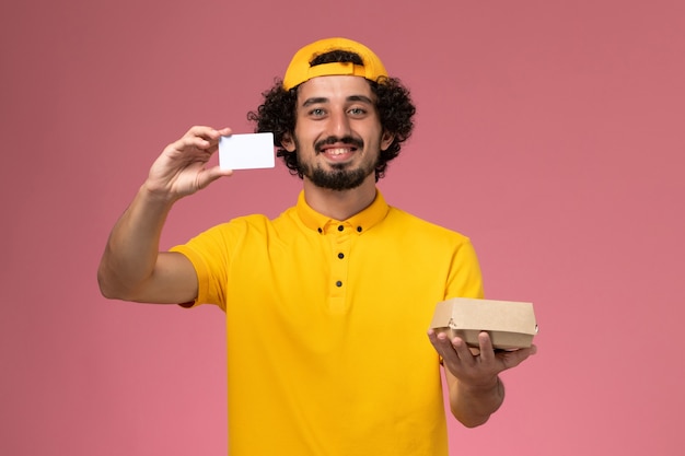 黄色のユニフォームとケープの正面図の男性の宅配便、薄ピンクの背景に彼の手にカードと小さな配達食品パッケージ。