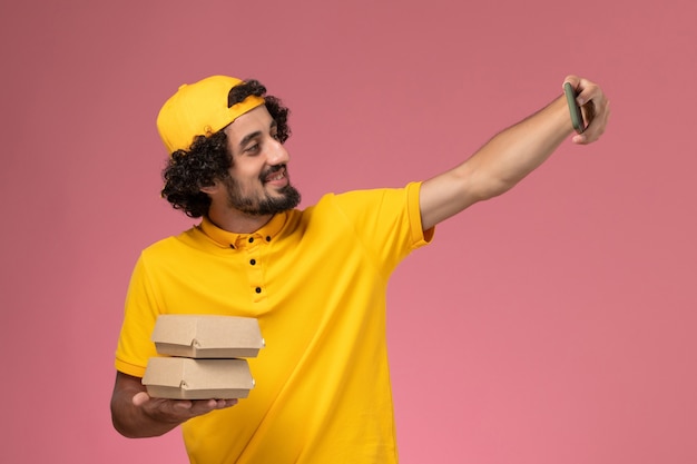 Foto gratuita corriere maschio vista frontale in mantello giallo uniforme con pacchi di cibo sulle mani che prendono foto su sfondo rosa chiaro.