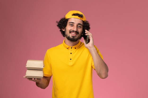Курьер-мужчина, вид спереди в желтой форме с пакетами с продуктами на руках, разговаривает по телефону на светло-розовом фоне.