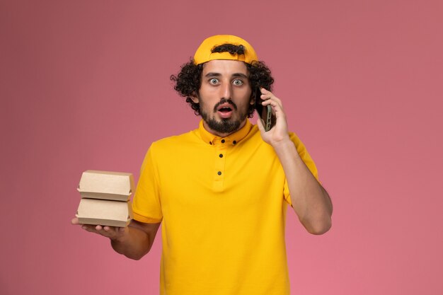 Front view male courier in yellow uniform cape with food packages on his hands talking on the phone on light-pink background.