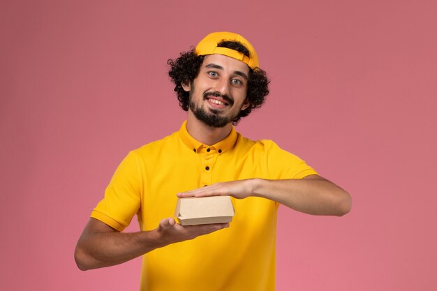 Front view male courier in yellow uniform and cape with little delivery food package on his hands on light-pink background.