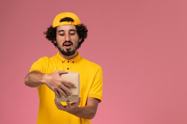 Front view male courier in yellow uniform and cape with little delivery food package on his hands opening it on the pink background.