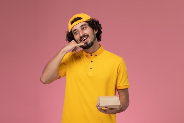 Front view male courier in yellow uniform and cape with little delivery food package on his hands on pink background.