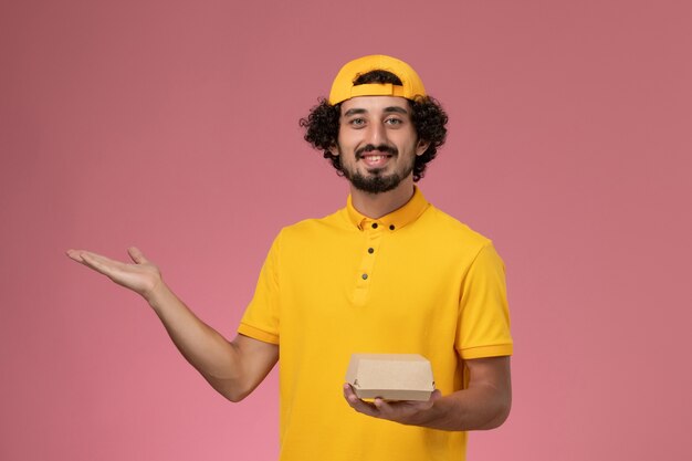 Front view male courier in yellow uniform and cape with little delivery food package on his hands on the pink background.