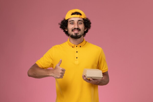 Курьер мужского пола вида спереди в желтой форме и плаще с небольшим пакетом еды доставки на его руках на розовом фоне.