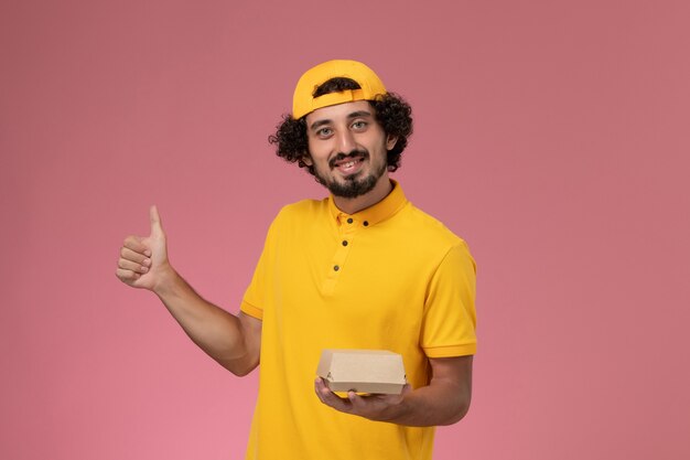 Front view male courier in yellow uniform and cape with little delivery food package on his hands on the pink background.