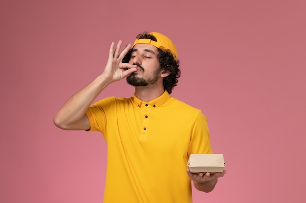 Free photo front view male courier in yellow uniform and cape with little delivery food package on his hands posing on the pink background.