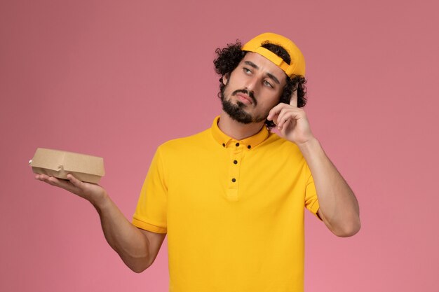 ピンクの背景を考えて彼の手に小さな配達食品パッケージと黄色の制服とケープの正面図の男性の宅配便。