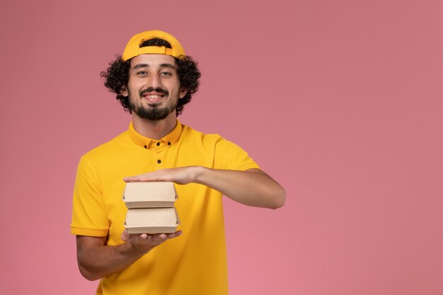 Front view male courier in yellow uniform and cape with little delivery food packages on his hands on the pink background.