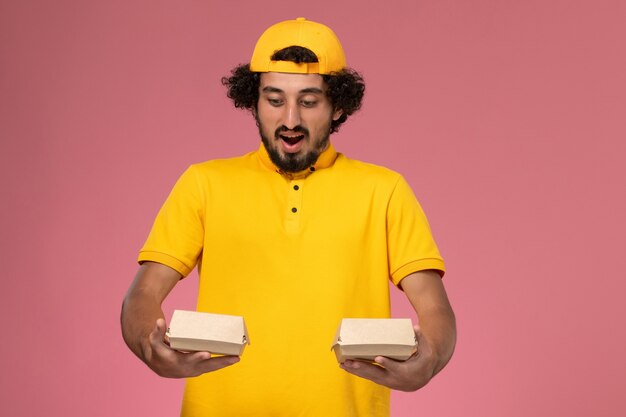 ピンクの背景に彼の手に小さな配達食品パッケージと黄色の制服とケープの正面図の男性の宅配便。