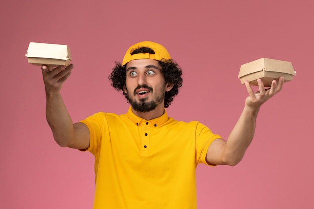 Foto gratuita corriere maschio vista frontale in uniforme gialla e mantello con piccoli pacchi di cibo di consegna sulle sue mani sullo sfondo rosa.