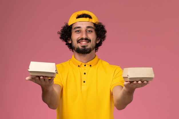 분홍색 배경에 그의 손에 작은 배달 음식 패키지와 노란색 유니폼과 케이프에서 전면보기 남성 택배.