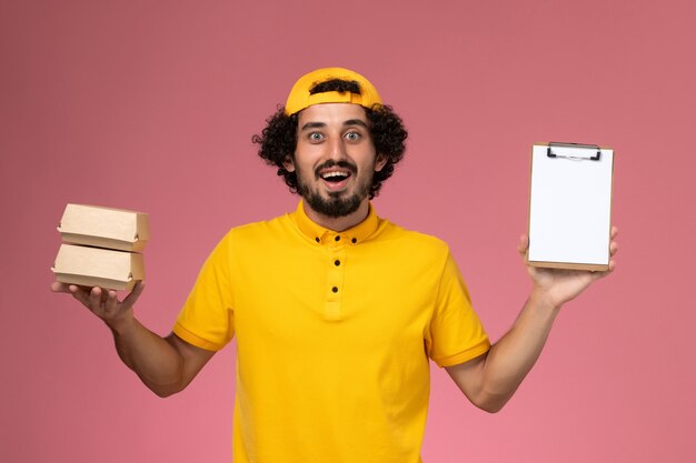 Front view male courier in yellow uniform and cape with little delivery food packages and notepad on his hands on the light pink background.