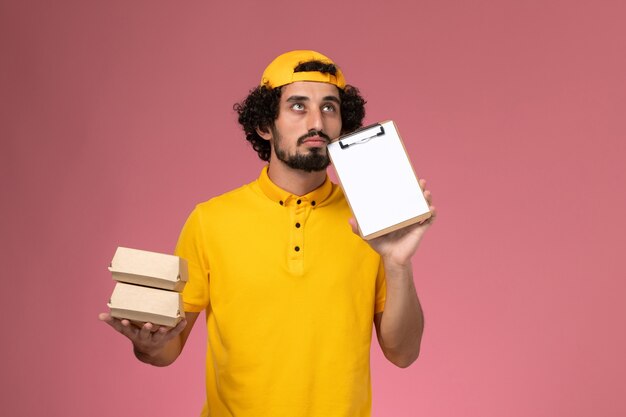 黄色のユニフォームとケープの正面図の男性の宅配便で、淡いピンクの背景に配達用の食品パッケージとメモ帳がほとんどありません。