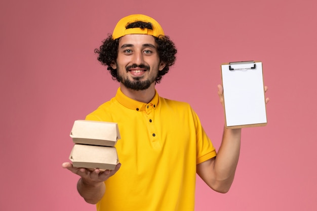 Front view male courier in yellow uniform and cape with little delivery food packages and notepad on his hands on the light pink background.