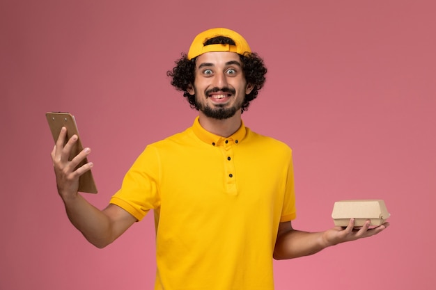 Front view male courier in yellow uniform and cape with notepad and little delivery food package on his hands on pink background.