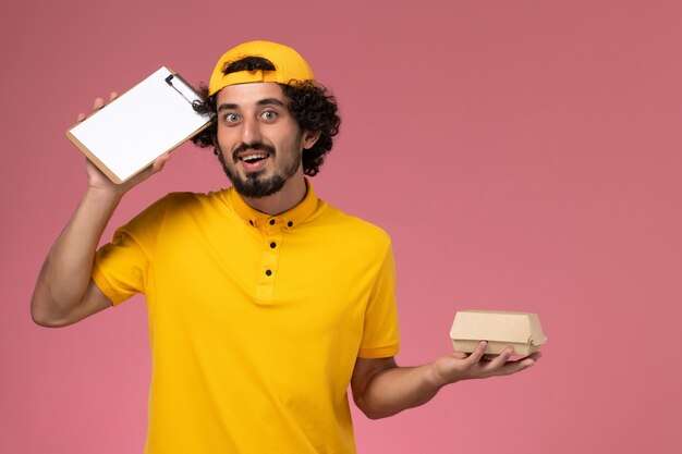 Front view male courier in yellow uniform and cape with notepad and little delivery food package on his hands on pink background.