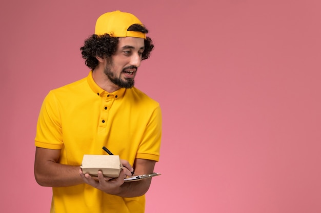 Front view male courier in yellow uniform and cape with notepad and little delivery food package on his hands and writing notes on pink background.