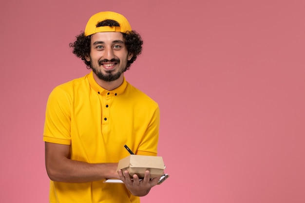 ピンクの背景にメモを書いている彼の手にメモ帳と小さな配達食品パッケージと黄色の制服とケープの正面図の男性の宅配便。