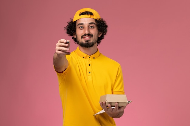 Front view male courier in yellow uniform and cape with notepad and little delivery food package pen on his hands on pink background.