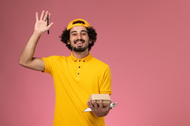 ピンクの背景に彼の手にメモ帳と小さな配達食品パッケージペンと黄色の制服とケープの正面図の男性の宅配便。