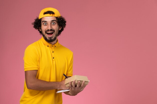 ピンクの背景に彼の手にメモ帳と小さな配達食品パッケージペンと黄色の制服とケープの正面図の男性の宅配便。