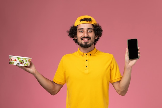 Front view male courier in yellow uniform cape with phone and delivery bowl on his hands on light-pink background.