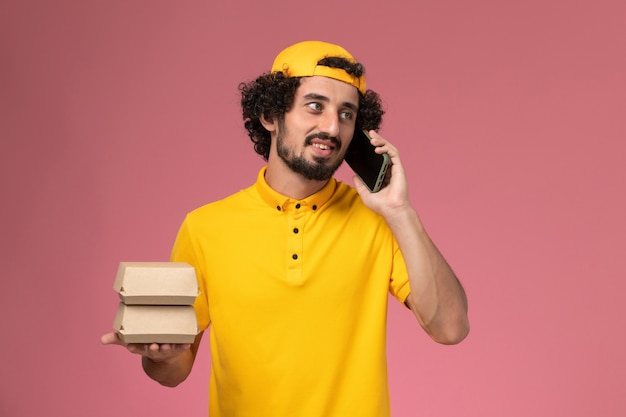 Front view male courier in yellow uniform cape with phone and food packages on his hands on light-pink background.