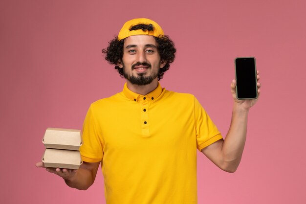 Front view male courier in yellow uniform cape with phone and food packages on his hands on light-pink background.