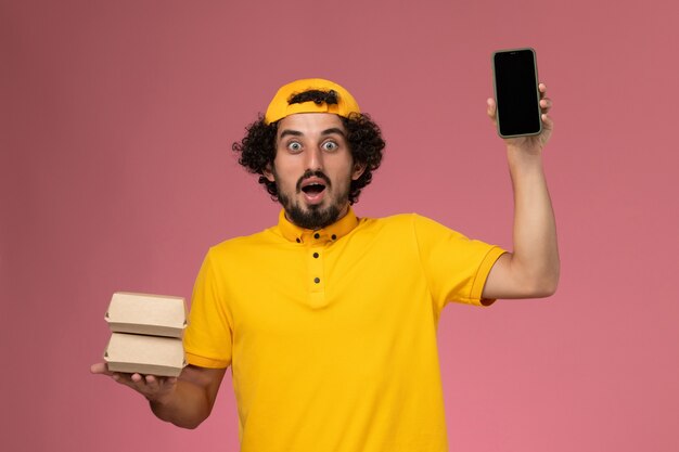 Front view male courier in yellow uniform cape with phone and food packages on his hands on light-pink background.