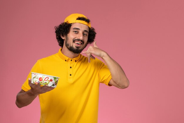 Front view male courier in yellow uniform and cape with round delivery bowl on his hands on the light pink background.