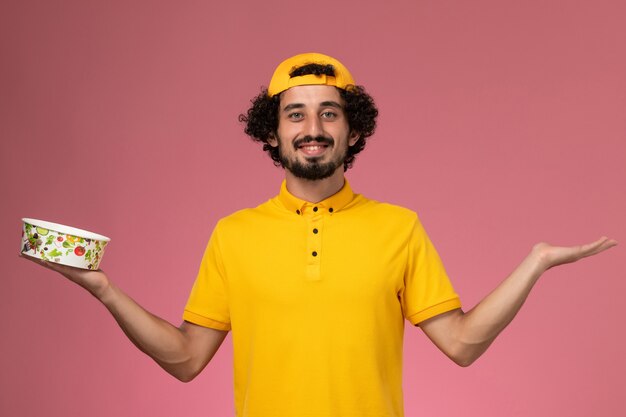 Front view male courier in yellow uniform and cape with round delivery bowl on his hands on the pink background.