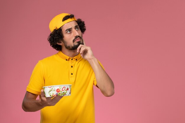 Front view male courier in yellow uniform and cape with round delivery bowl on his hands on the pink background.