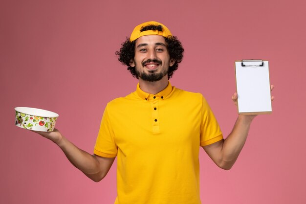 Front view male courier in yellow uniform cape with round delivery bowl on his hands on the pink background.