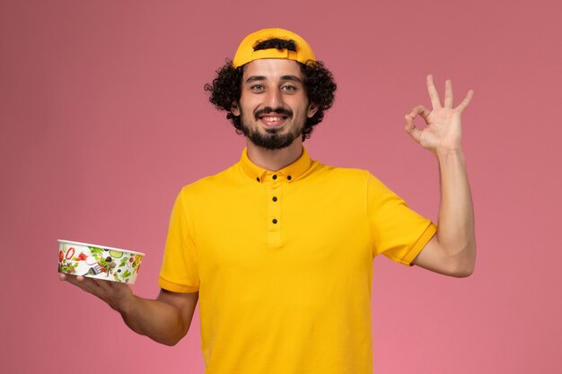 Front view male courier in yellow uniform and cape with round delivery bowl on his hands smiling on the pink background.