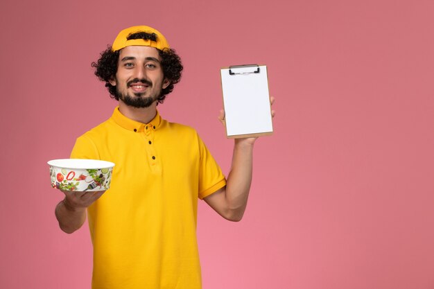 Front view male courier in yellow uniform cape with round delivery bowl on his hands and with notepad on the pink background.