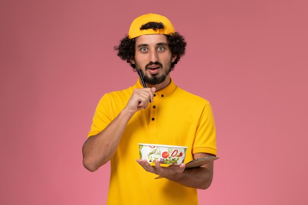 Front view male courier in yellow uniform cape with round delivery bowl on his hands writing notes on the pink background.