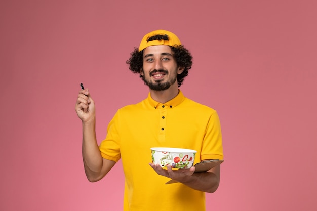 Free photo front view male courier in yellow uniform cape with round delivery bowl on his hands writing notes on the pink background.