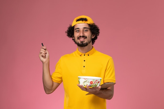 분홍색 배경에 메모를 작성하는 그의 손에 둥근 배달 그릇으로 노란색 유니폼 케이프에서 전면보기 남성 택배.