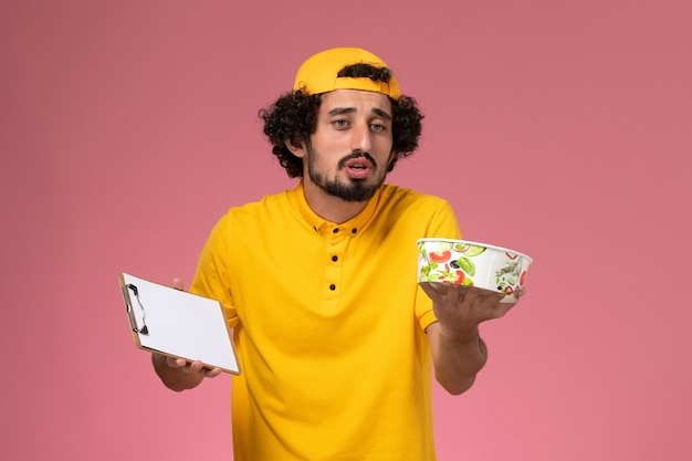 Front view male courier in yellow uniform cape with round delivery bowl and notepad on his hands on light-pink background.