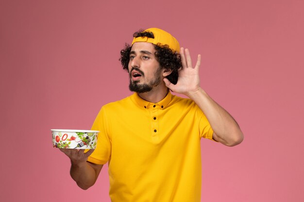 Front view male courier in yellow uniform and cape with round delivery bowl trying to hear on his hands on the light pink background.