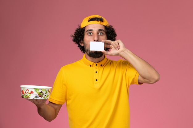 Front view male courier in yellow uniform cape with round delivery bowl with white card on his hands on the pink background.