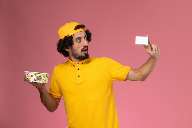 ピンクの背景に彼の手に白いカードと丸い配達ボウルと黄色の制服ケープの正面図男性宅配便。