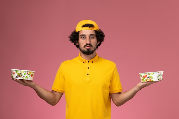 Front view male courier in yellow uniform and cape with round delivery bowls on his hands on the light pink background.