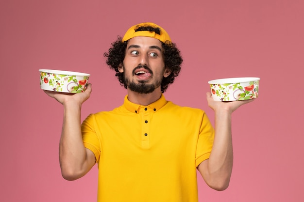 Free photo front view male courier in yellow uniform and cape with round delivery bowls on his hands on the light pink background.