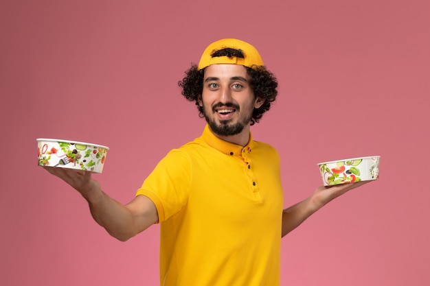 Front view male courier in yellow uniform and cape with round delivery bowls on his hands on the light pink background.