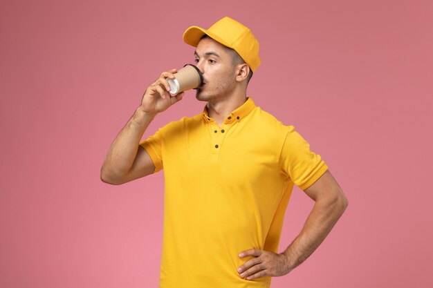 Front view male courier in yellow uniform drinking coffee on the pink background 